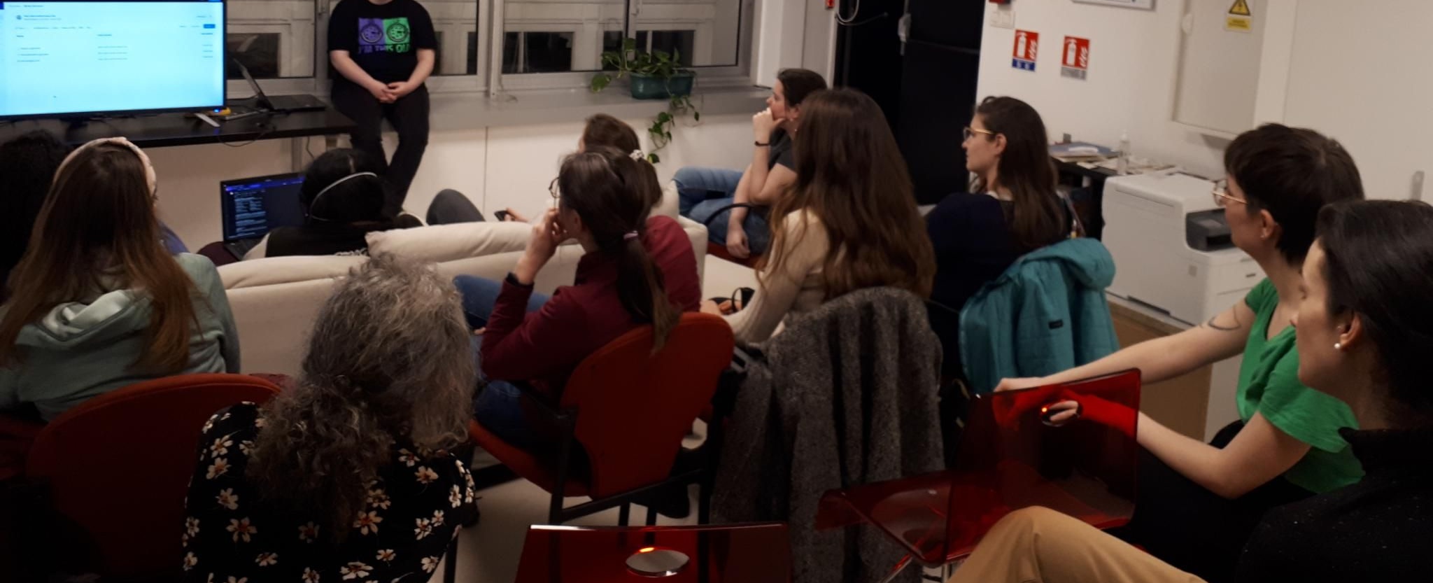 une dizaine de femmes de dos, assises sur des sièges dépareillés, regardent une présentation donnée également par une femme. On aperçoit une télévision sur laquelle est projetée la présentation.